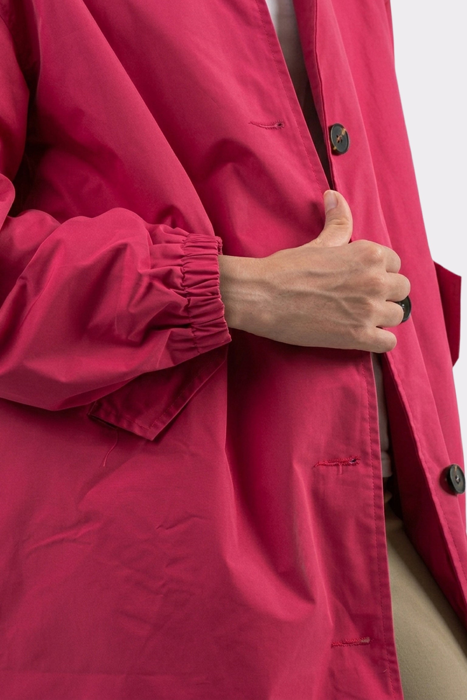 A person holds the front of The Water Resistant Hooded Oversized Windbreaker Rain Jacket, featuring bright pink fabric, black buttons, an elastic cuff on the sleeve, and a beige garment visible underneath.