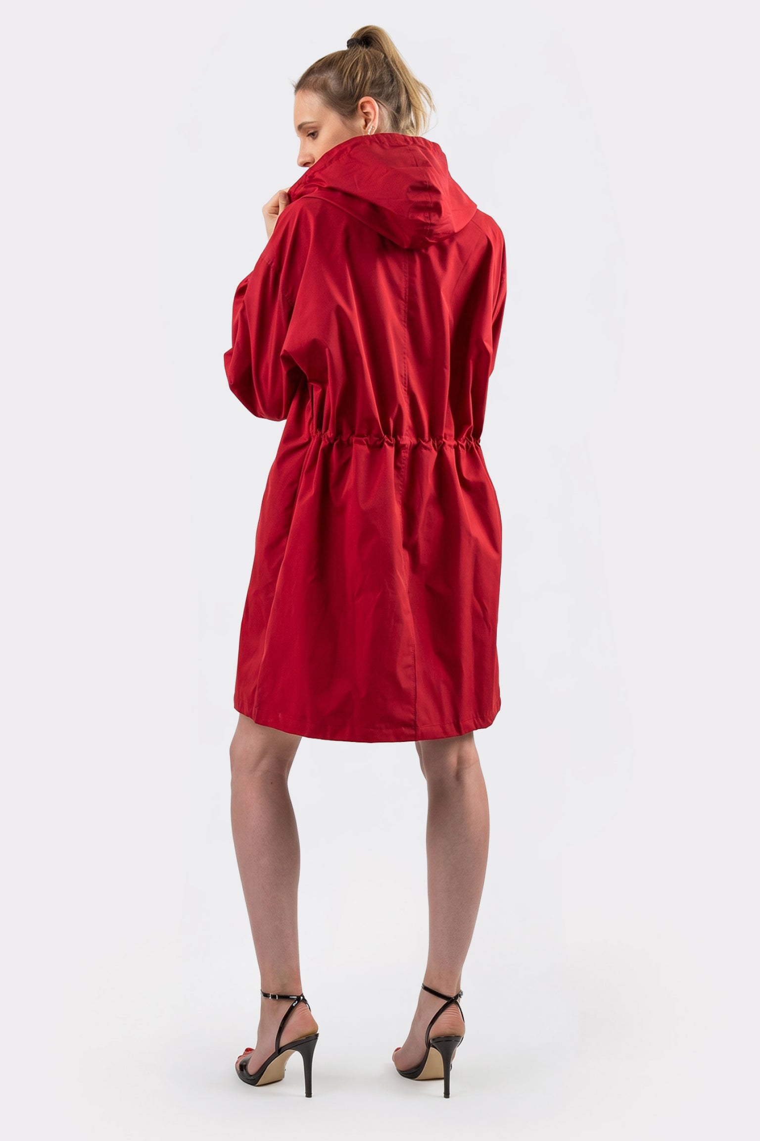 A person faces away, wearing The Water Resistant Hooded Oversized Windbreaker Rain Jacket in red with an elastic waist and black high-heeled sandals, set against a plain, light background.