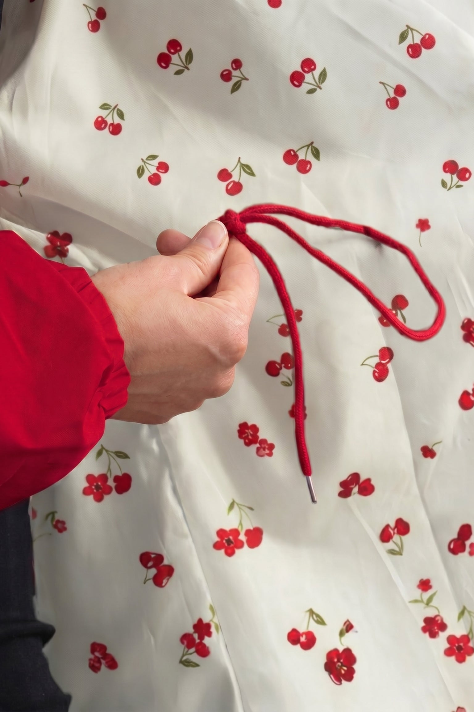 Someone in a red-sleeved top holds the drawstring of The Water Resistant Hooded Oversized Windbreaker Rain Jacket, shown in white with a red cherry and green leaf print.