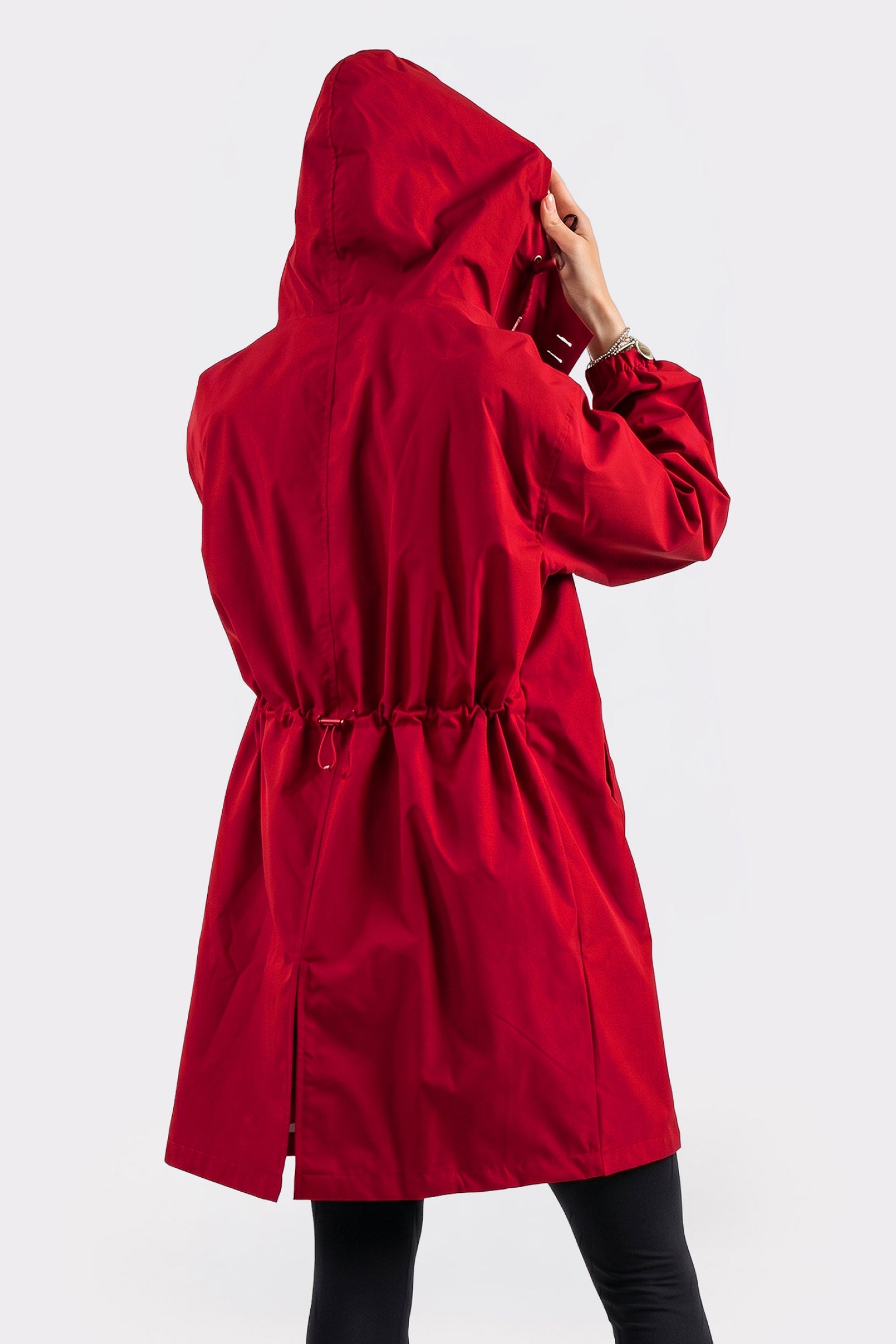 A person wearing The Water Resistant Hooded Oversized Windbreaker Rain Jacket in red stands with their back to the camera, adjusting the hood. The jacket features a drawstring waist and is set against a plain white background.