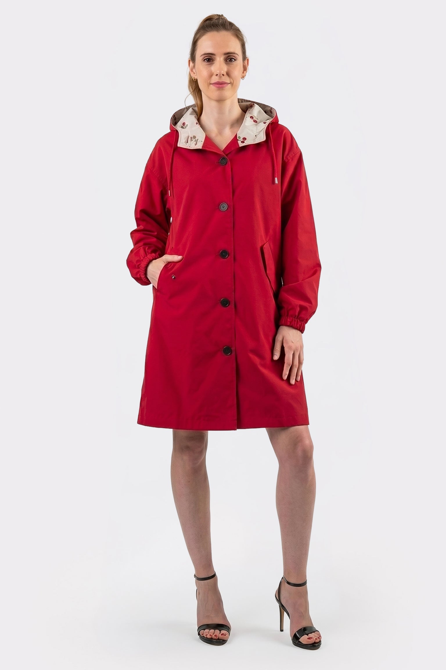 A person stands facing forward in The Water Resistant Hooded Oversized Windbreaker Rain Jacket, a knee-length red coat with black buttons and a beige patterned hood, paired with black high-heeled sandals. One hand is in a pocket; background is plain white.