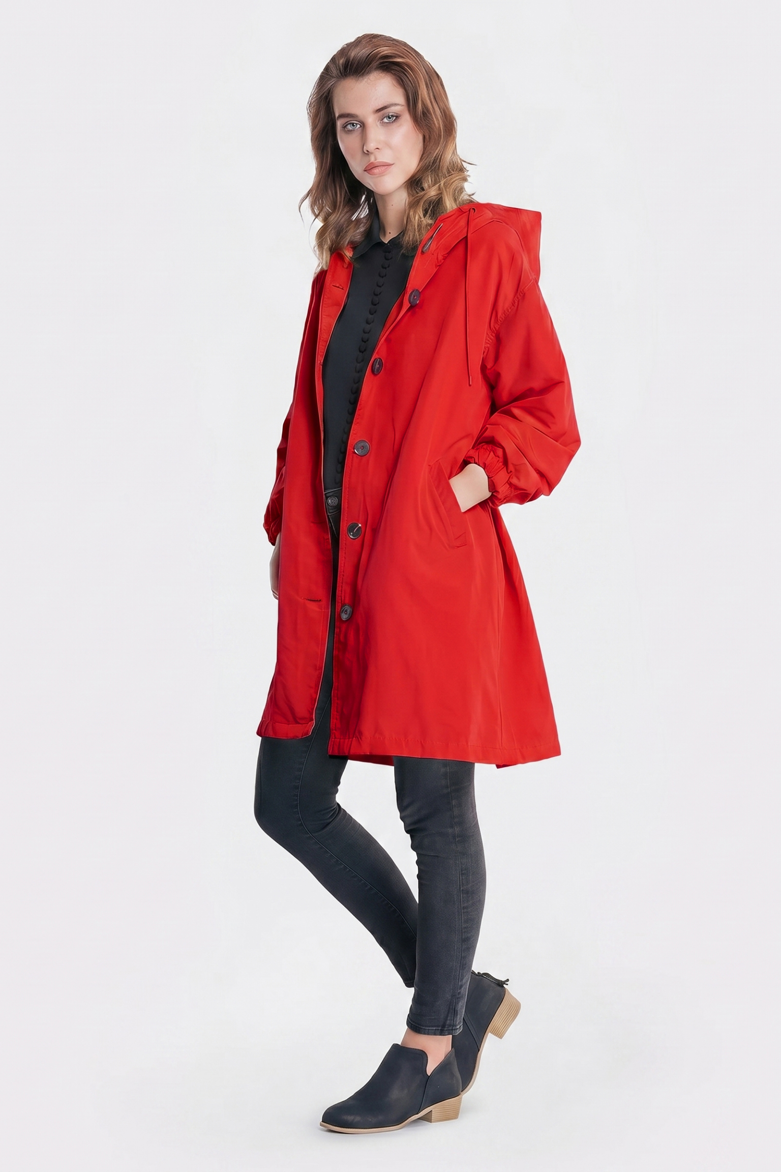 A person poses against a plain white background wearing the Icon Windbreaker Rain Jacket in bright red with a hood, paired with a black shirt, black skinny jeans, and black low-heeled slip-on shoes.