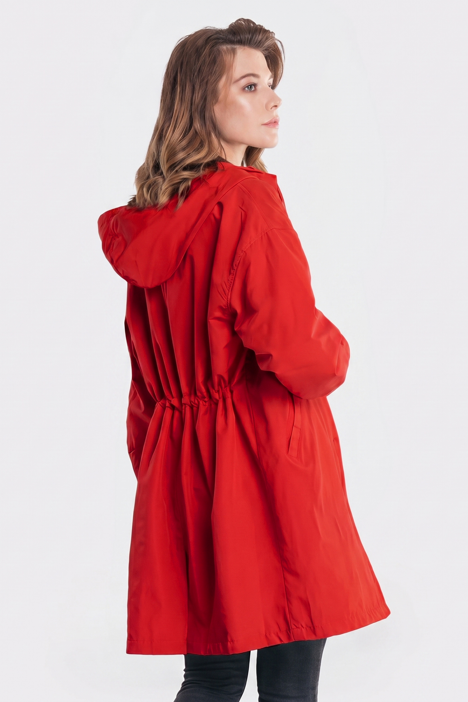 A woman with light brown hair wears the Icon Windbreaker Rain Jacket in red, hands in pockets, coat gathered at the waist, standing at an angle against a plain white background.