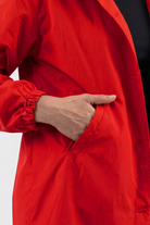 A person wears a bright red Icon Windbreaker Rain Jacket with elastic cuffs, placing their right hand in the pocket. The background is plain, and they have a black garment underneath.