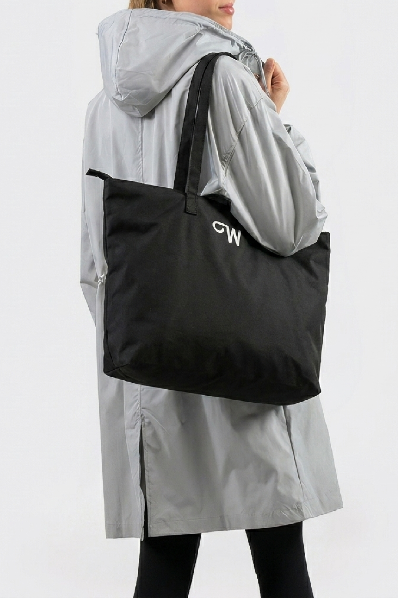 A person in a light gray hooded coat and black leggings holds the large black Urban Slate Waterproof Bag with "CW" printed on it, standing against a plain, light-colored background.