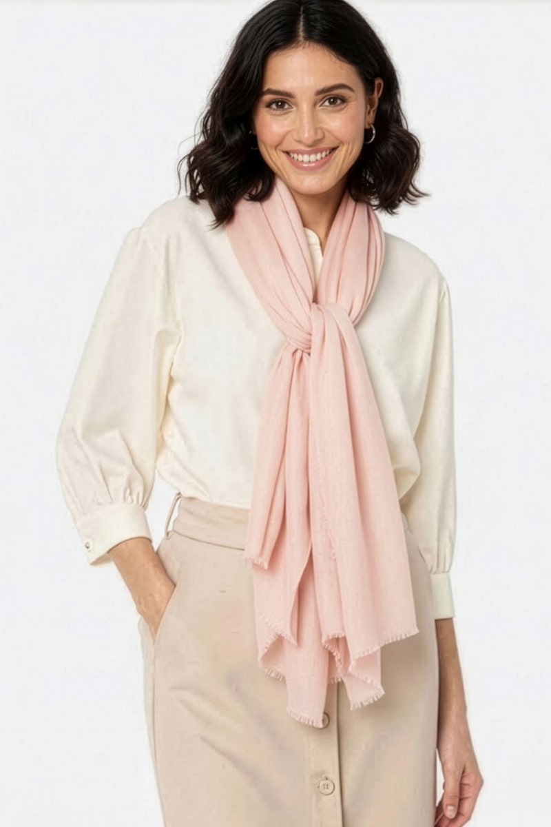 A woman with shoulder-length dark hair, wearing a cream blouse, beige skirt, and the Blush Rose 100% Cashmere Scarf by looselyboho loosely tied around her neck, stands against a plain background and smiles at the camera.