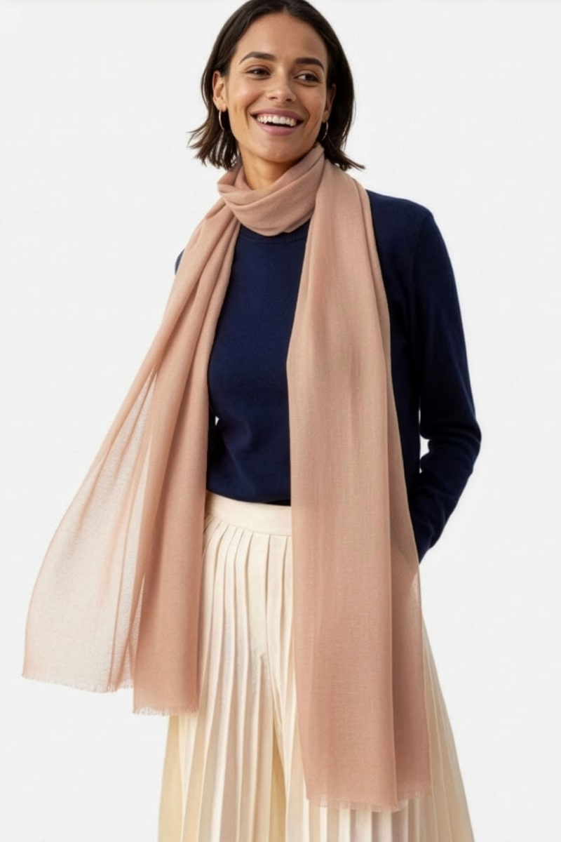 A woman stands against a light background, smiling and looking to the side. She wears a navy long-sleeve top, a pleated cream skirt, and the looselyboho Sandstone Dune 100% Cashmere Scarf draped effortlessly around her neck.