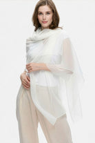A woman with light brown hair and a slight smile stands against a plain background, elegantly draped in the looselyboho Ivory Whisper 100% Cashmere Scarf over beige pants and a white top, capturing minimalist luxury.