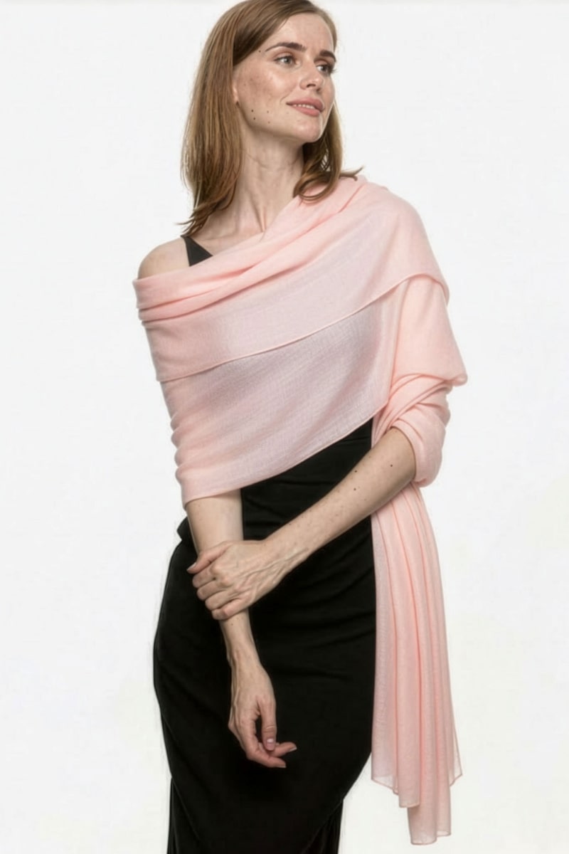 A woman with light brown hair wears a sleeveless black dress and the looselyboho Blush Rose 100% Cashmere Scarf, elegantly draped over her shoulders, standing against a plain white background.