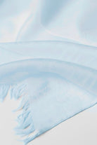 A close-up of the looselyboho Sky Mist 100% Cashmere Scarf, featuring a soft, semi-sheer light blue fabric with fringed edges, displayed on a white surface.