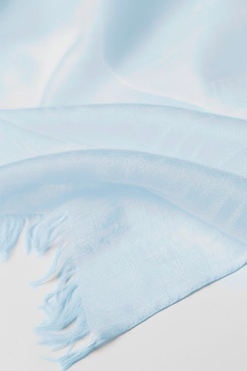 A close-up of the looselyboho Sky Mist 100% Cashmere Scarf, featuring a soft, semi-sheer light blue fabric with fringed edges, displayed on a white surface.