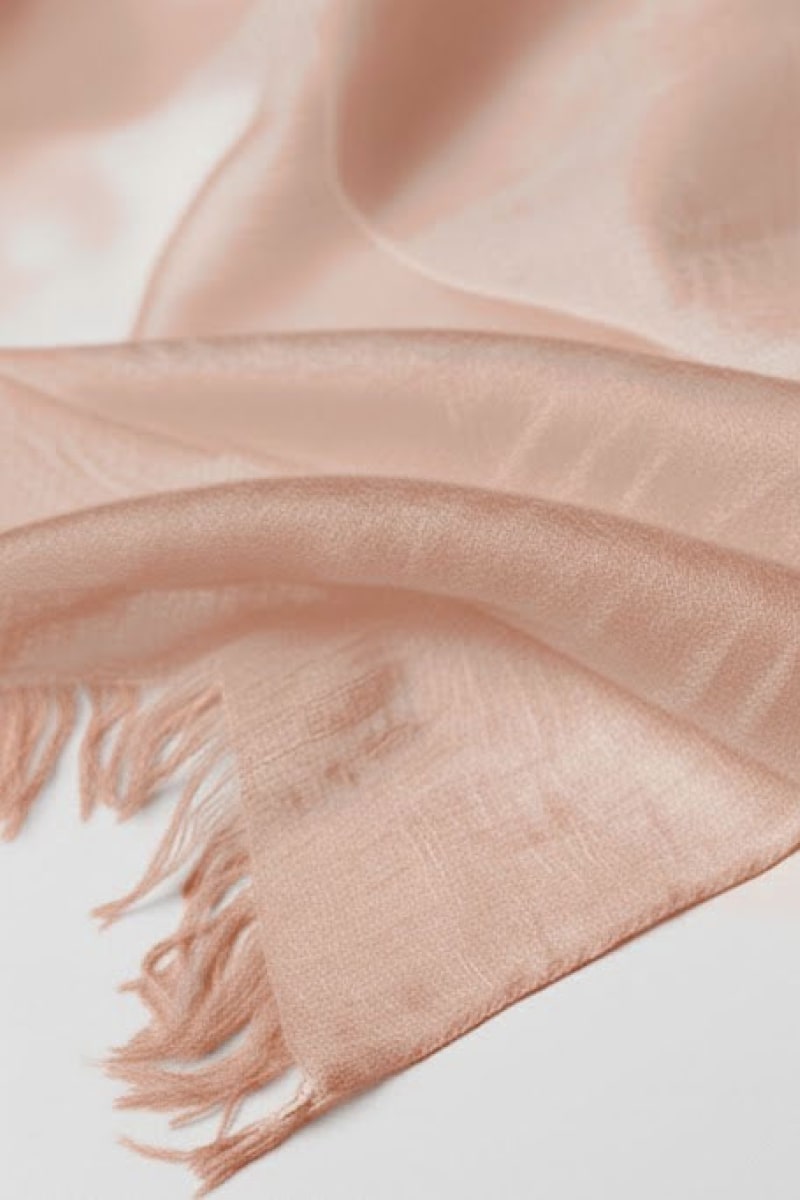 A close-up of the looselyboho Sandstone Dune 100% Cashmere Scarf in light pink shows its semi-sheer, fringed fabric with a smooth texture and wispy edges—perfect for elegant winter layering.