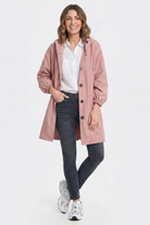 A woman smiles while wearing the Icon Windbreaker Rain Jacket in light pink, featuring a hood, paired with a white collared shirt, dark skinny jeans, and white sneakers. She stands against a plain white background with one hand in her pocket.