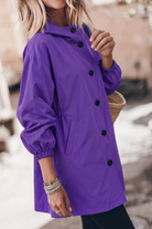 A person with shoulder-length blonde hair wears a loose-fitting, long-sleeve purple Icon Rain Windbreaker Jacket with black buttons. Their face is out of frame. They wear metal bangles and hold a woven bag. The background is blurred.