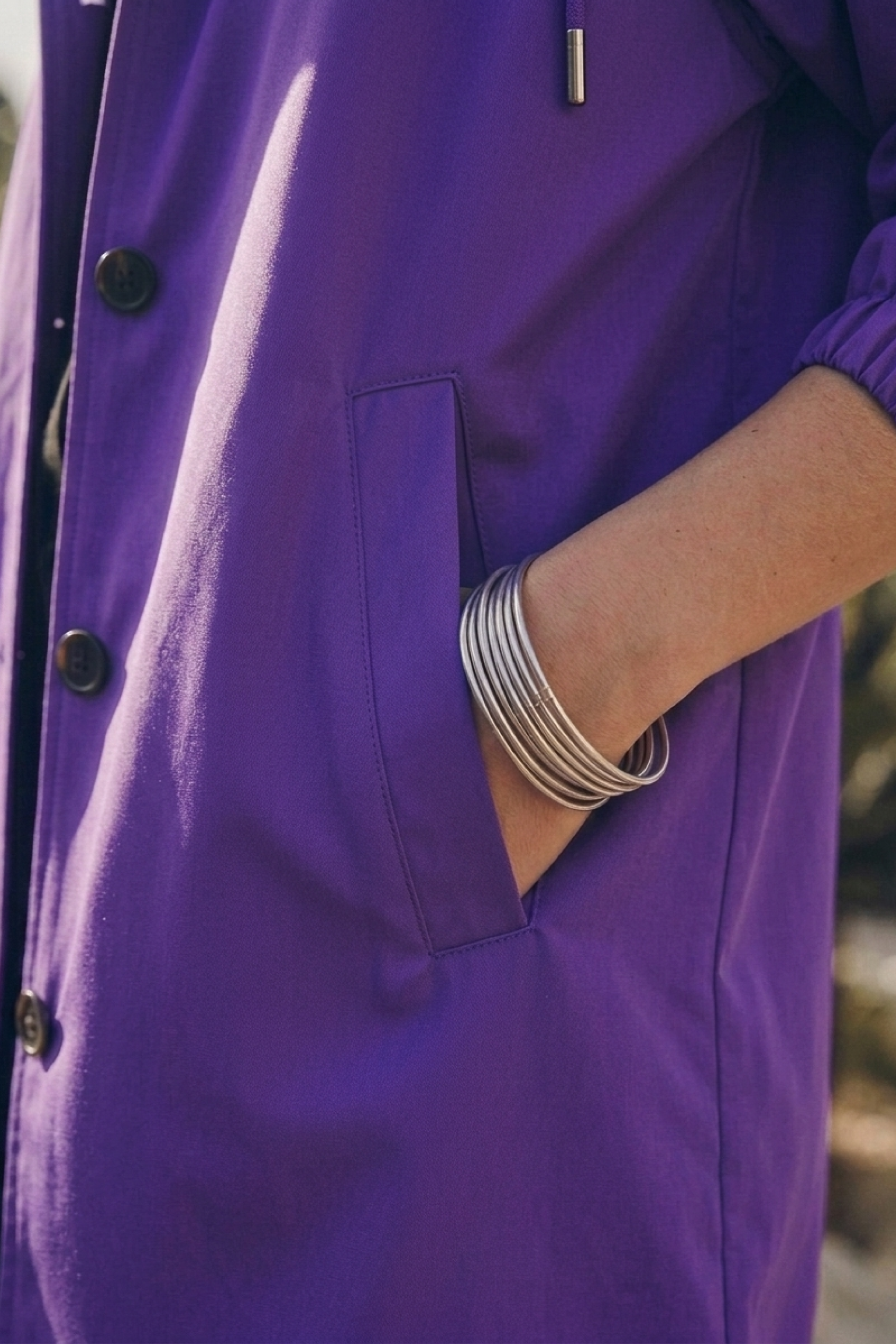 A person wearing the Icon Rain Windbreaker Jacket in purple, featuring black buttons, has their right hand tucked into the pocket. Several silver bangles adorn their wrist. The water-resistant jacket stands out vividly against a slightly blurred background.