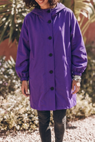 A person stands outdoors wearing the Icon Rain Windbreaker Jacket—a loose-fitting, long-sleeved purple rain jacket with black buttons—paired with dark pants and bracelets. The background features plants and gravel, and the person’s face is partially out of frame.