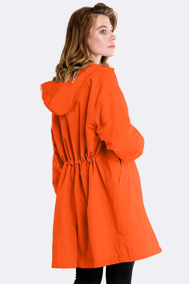 A person wearing the looselyboho PUMPKIN ORANGE WINDBREAKER, featuring a cinched waist, stands facing left and glances over their shoulder. With the hood down, their long hair is visible against a plain white background.