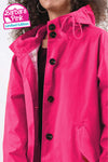 Individual wearing a bright pink raincoat with black buttons and a hood. A label on the top left corner reads "looselyboho BARBARA PINK WINDBREAKER." Made from lightweight water-resistant fabric, this looselyboho BARBARA PINK WINDBREAKER reveals a maroon shirt partially visible underneath. The person has curly hair.