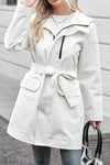 A woman in a white looselyboho BELTED FROST WHITE WINDBREAKER stands outdoors. The coat has a high collar and large front pockets. She holds a black handbag and wears a white top underneath. The setting appears to be an urban sidewalk with a grey building wall in the background.