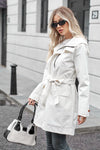 A person with long blonde hair wears a stylish BELTED FROST WHITE WINDBREAKER by looselyboho and dark sunglasses. They are carrying a black and white handbag and standing in front of a building with large windows and a door marked with the number 29.