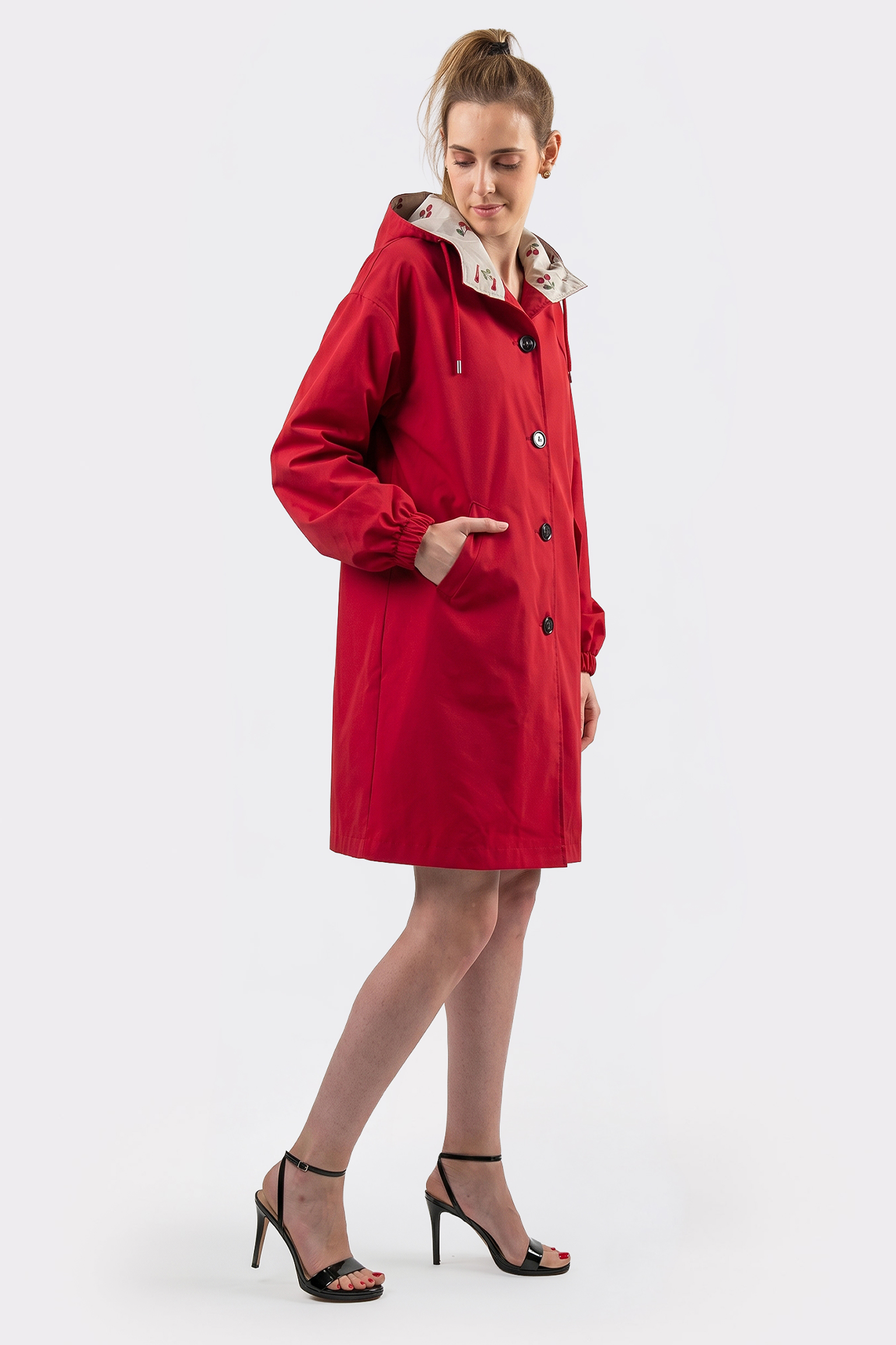 A person wears The Water Resistant Hooded Oversized Windbreaker Rain Jacket in red with black buttons, hands in pockets, looking down and to the side. They pair it with black high-heeled sandals against a plain white background.