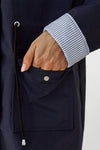 A person wearing a STRIPED NAVY SHADOW WINDBREAKER JACKET by looselyboho is shown with their left hand in the jacket's pocket. The pocket has a buttoned flap, and a black drawstring with a silver end is visible near the pocket.