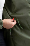 A person with a red manicure adjusts the drawstring on an olive green, lightweight water-resistant OLIVE GREEN WINDBREAKER from The Windbreaker Jacket. Only the hand, part of the arm, and the lower torso are visible. They are wearing a white shirt and dark pants.