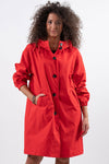 A person with curly hair stands against a white background, wearing a stylish CRIMSON RED WINDBREAKER by looselyboho with large black buttons and two pockets. The person is smiling slightly and has one hand in a pocket.