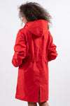 A person with curly hair is standing with their back to the camera, wearing a stylish CRIMSON RED WINDBREAKER by looselyboho. The jacket features a waist cinch, a rear slit at the bottom, and offers water-resistant protection. The background is a plain, light gray.