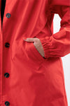 Close-up of a person wearing a stylish CRIMSON RED WINDBREAKER by looselyboho in bright red, with their right hand in the coat's pocket. The coat has black buttons and the sleeve features an elastic cuff. The background is white and the person has red nail polish matching the water-resistant jacket.